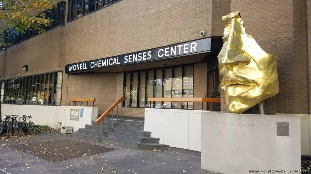 A tan building with gray steps leading up to an entrance bordered by white walls. The words Monell Chemical Senses Center is on a sign across the front of the building. A golden statue of a face stands to one side with a plague underneath.